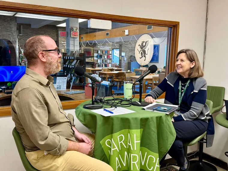 Two people are sitting at a round table with microphones, in a room with a glass window showing a library in the background.