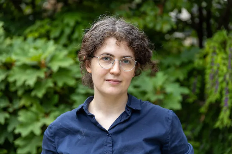 Person with short curly hair and glasses in a dark shirt, standing outdoors in front of leafy greenery.