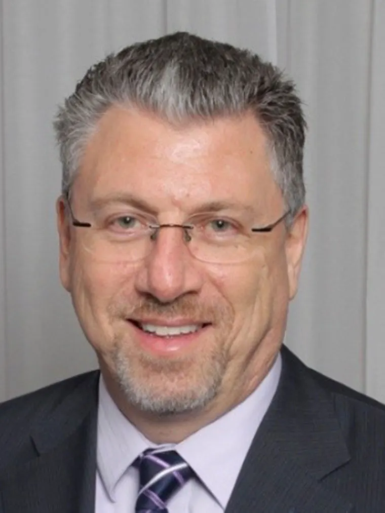 Person with short gray hair, glasses, a suit, and a patterned tie, smiling at the camera against a plain curtain background.