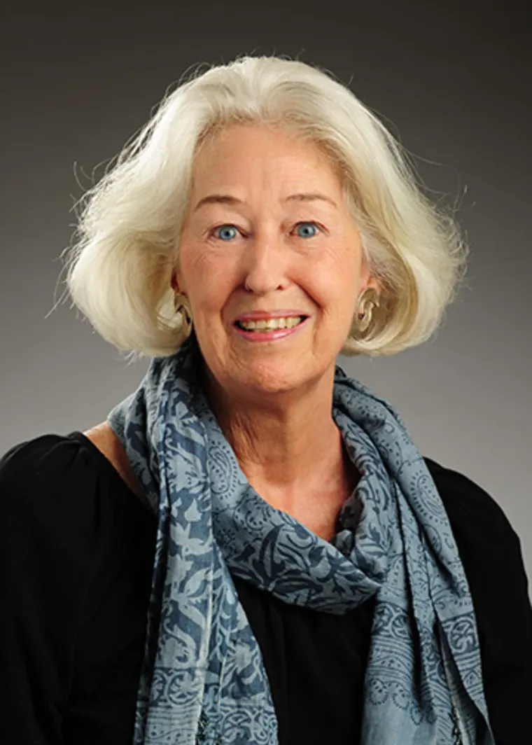 Person with short white hair, wearing a blue patterned scarf and black top, smiling against a neutral background.
