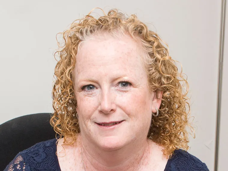 Person with curly hair and light complexion, smiling slightly, wearing a blue top, in front of a plain background.