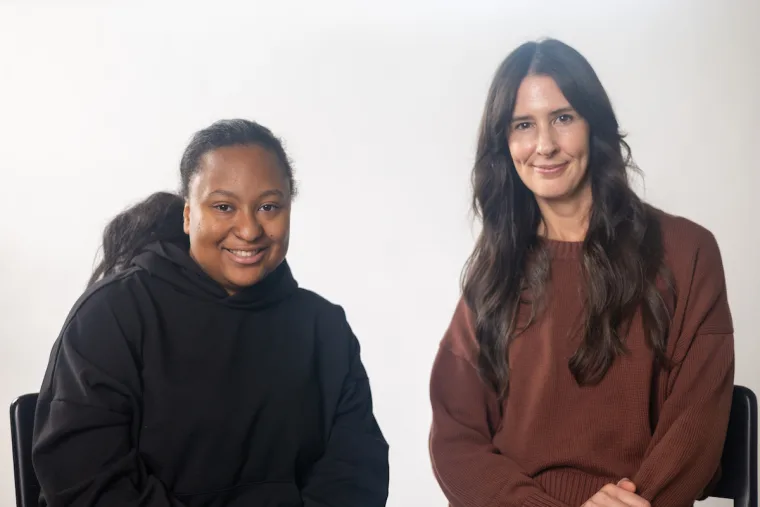Two people sit side by side, both smiling. One wears a black hoodie, the other a brown sweater. The background is plain white.