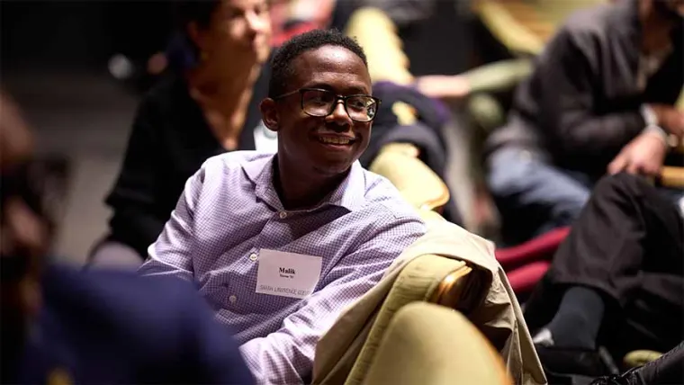 A person wearing glasses and a name tag is seated, looking to the left, with others sitting nearby in a dimly lit room.
