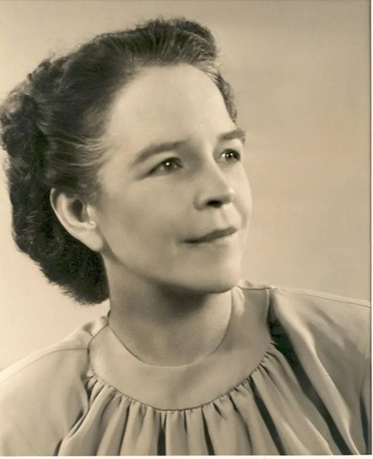 Portrait of a person looking upwards with a neutral expression, wearing a gathered neckline blouse, and hair styled back.