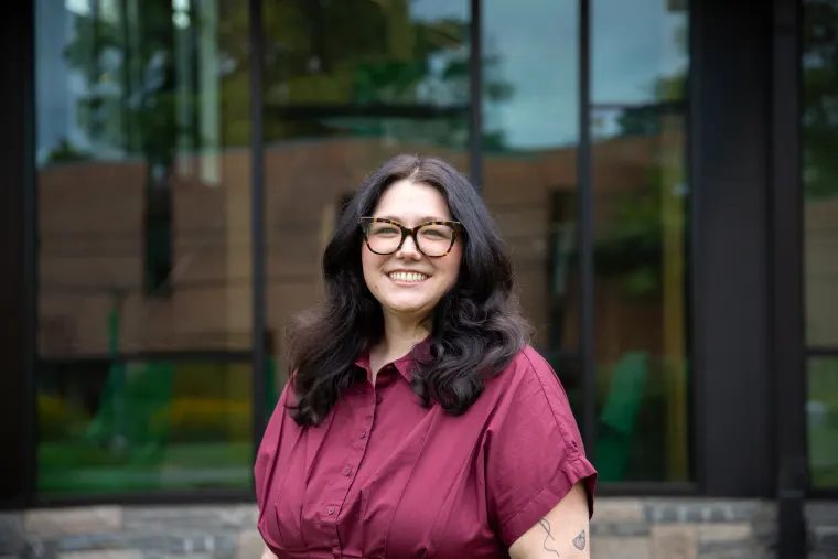 Person with long dark hair and glasses smiling, wearing a maroon shirt, standing in front of a glass building.