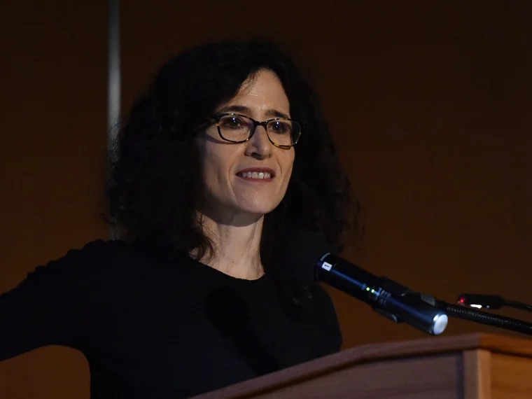 Person with curly hair and glasses speaking at a podium with a microphone, background dimly lit.