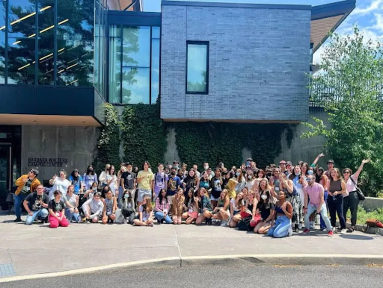A large group of people posing together in front of a modern building with ivy-covered walls and trees around, on a sunny day.