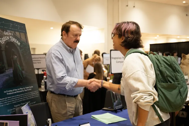 Two people shaking hands at a table with promotional material in a busy indoor event setting.