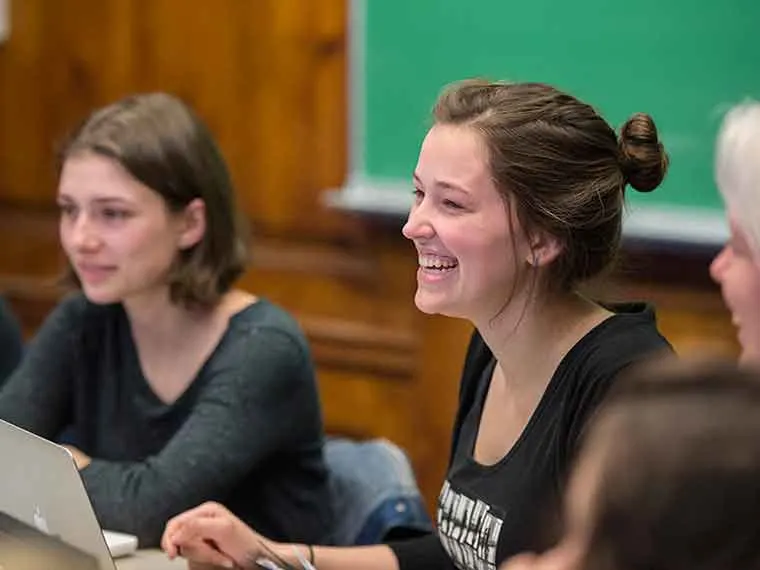 People sitting at a table in a classroom, one smiling, with a chalkboard in the background.