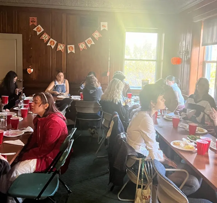 People gather around tables in a room with wooden walls and autumn-themed decorations, talking and eating with red cups and plates.