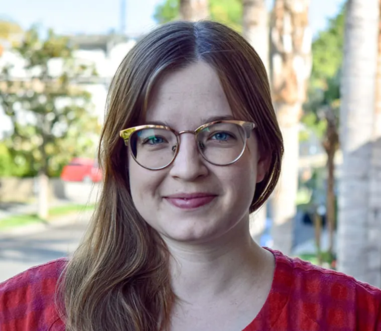 Person with long hair and glasses smiling outdoors, wearing a red top. Trees and buildings visible in the background.
