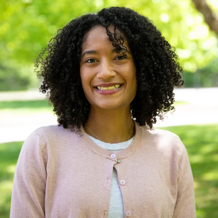 Person smiling outdoors with curly hair, wearing a pink cardigan and standing in front of green trees in a sunny park setting.