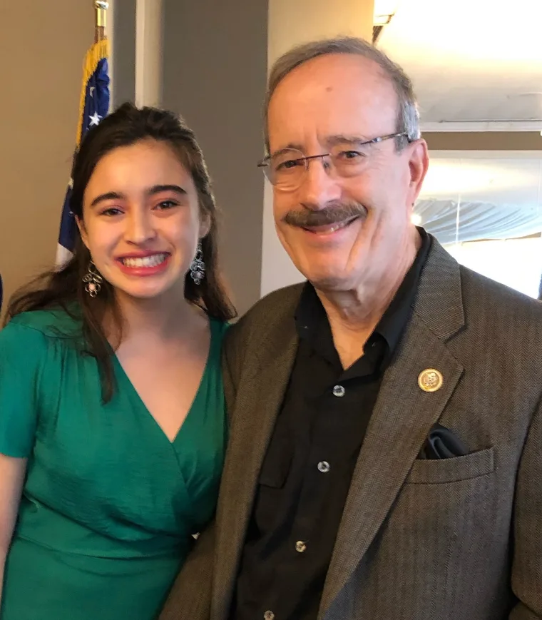 Two people smiling, standing close together indoors, one is wearing a green dress, the other a gray blazer with a round pin, with a flag partially visible.