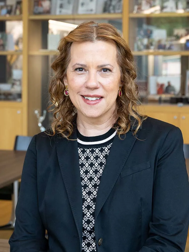 Person with curly hair smiling, wearing a black blazer with a patterned shirt, in an office with shelves in the background.