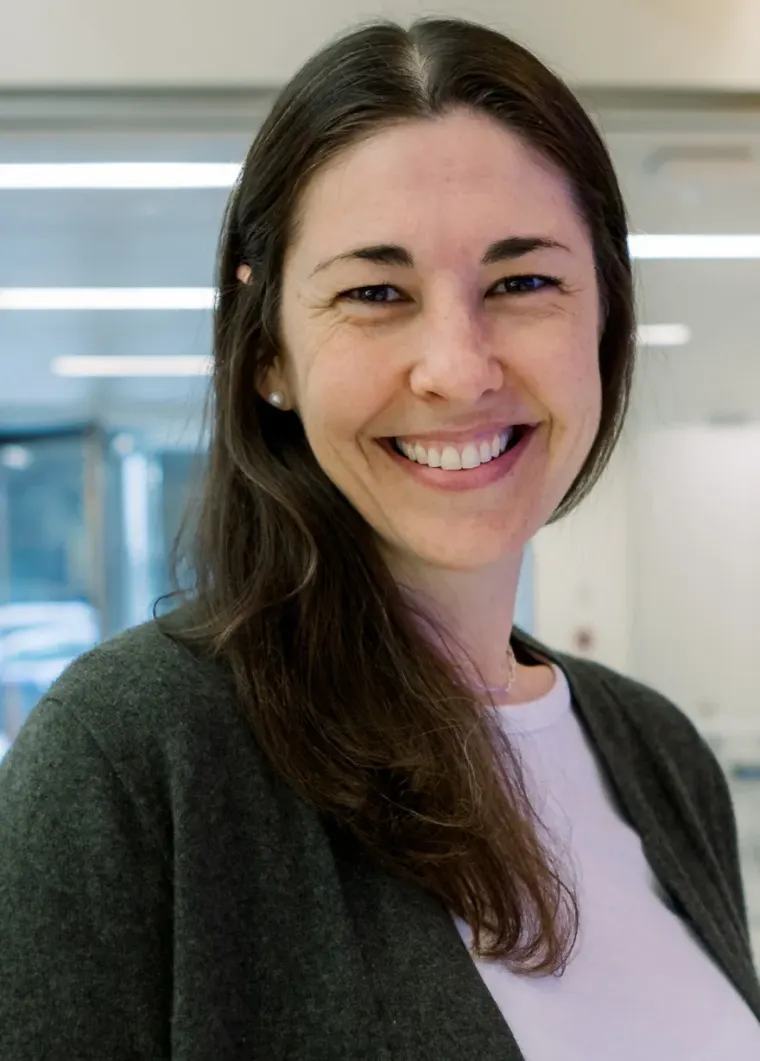 Person with long dark hair, smiling, wearing a gray cardigan and white top in a bright indoor setting.