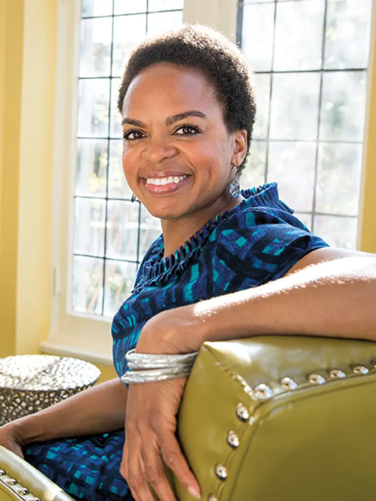 Person smiling while sitting on an olive green chair, wearing a blue patterned shirt and silver bangles, with large windows in the background.
