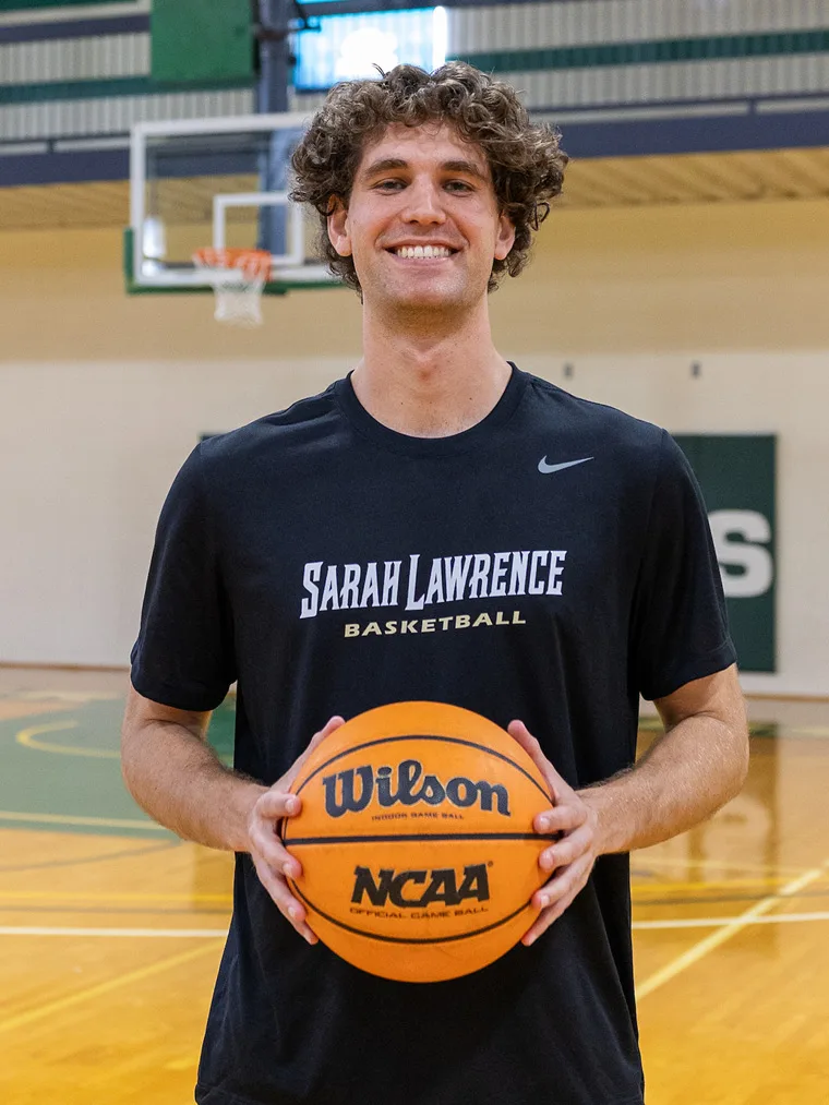 Person with curly hair holds a basketball on an indoor court, wearing a "Sarah Lawrence Basketball" shirt.