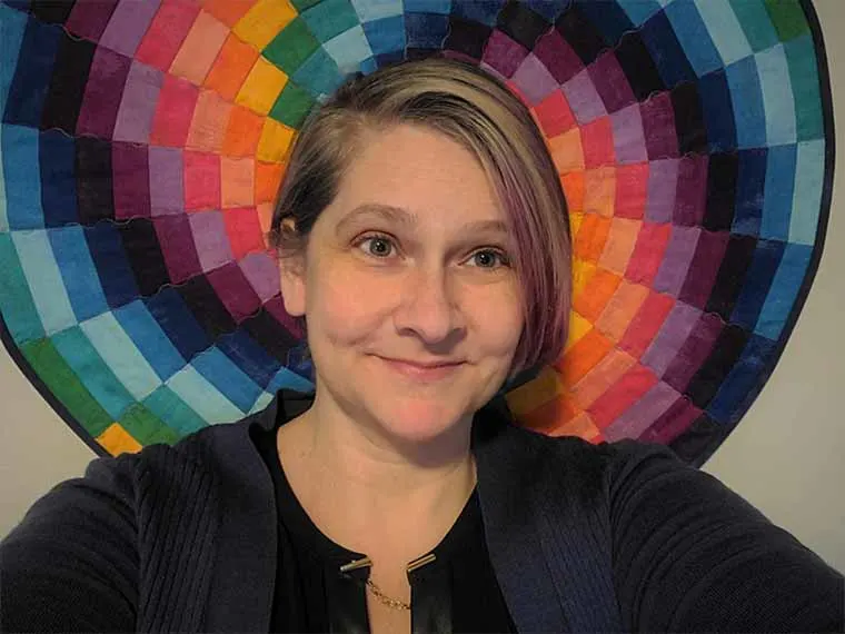 Person smiling in front of a circular, multicolored quilt with a pattern resembling a rainbow.