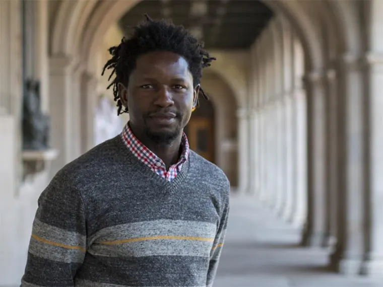 Person with short dreadlocks in a sweater and checkered shirt stands in a colonnade with arched ceilings and blurred background.