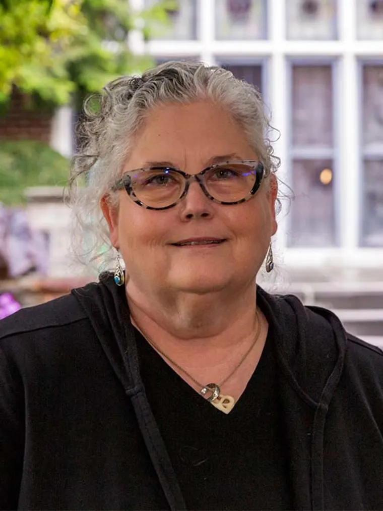 Person with glasses and curly hair wearing a black hoodie and necklace, standing in front of a windowed background with greenery.