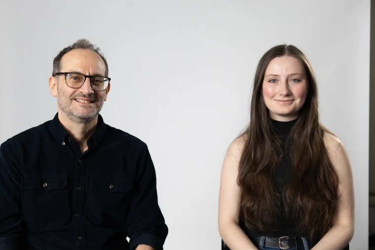 Two people sitting side by side against a plain white background, one wearing glasses and a dark shirt, and the other with long hair.