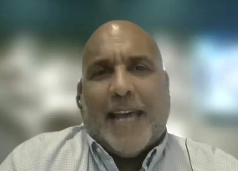 Person speaking on a video call, wearing a light-colored collared shirt with a blurred background.