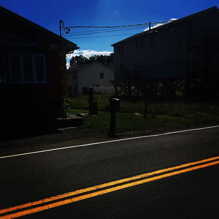 Road with double yellow lines. Two darkened houses in the background, one on stilts. Bright sky with faint clouds.