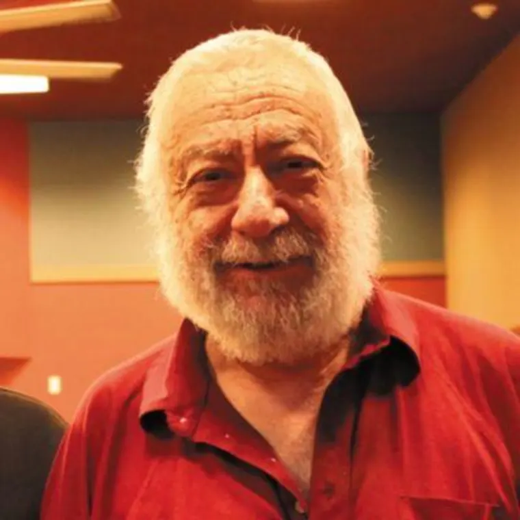 Person with a white beard, smiling, wearing a red shirt, stands indoors against a warm-colored background.