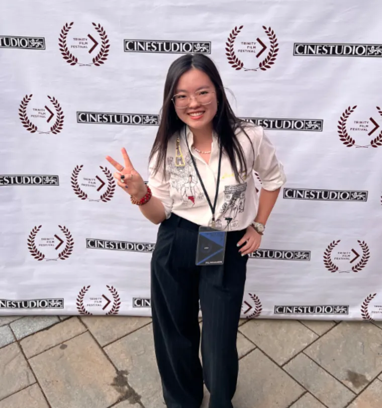 Person smiling and holding up a peace sign in front of a backdrop with "Cinestudio" and "Trinity Film Festival" logos.