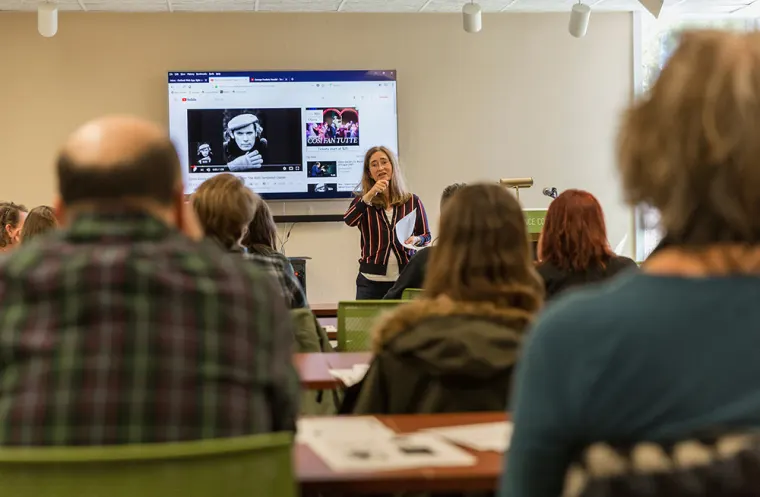 Person speaking in a classroom, holding papers, with a display showing videos behind. Audience seated with backs facing the camera.