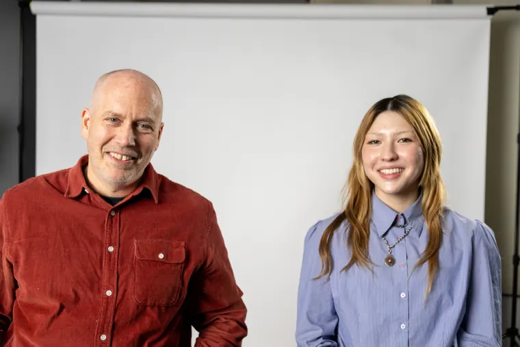 Two people smiling in front of a photography backdrop; one wears a red shirt, the other a blue shirt with a necklace.