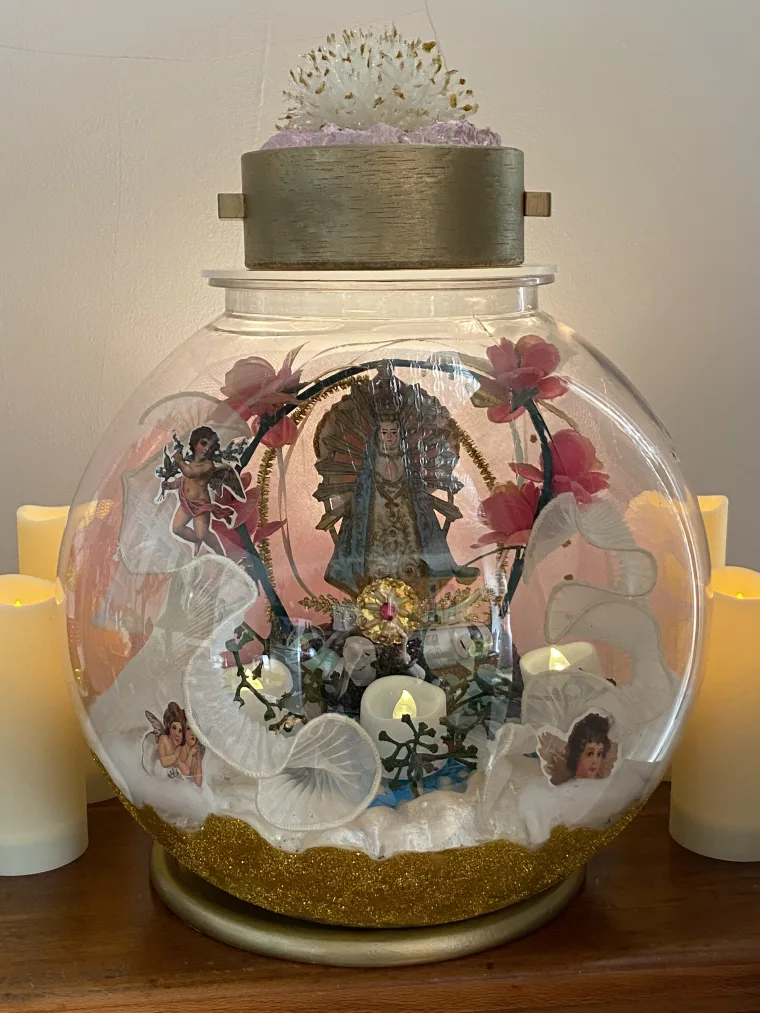 Decorative glass dome with religious figure, flowers, and angel figurines. Surrounded by candles, with a textured object on top.