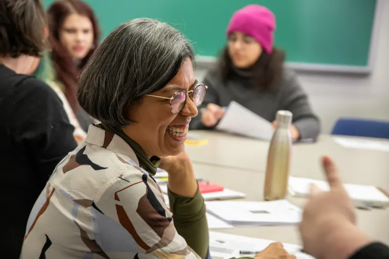 Person with glasses smiling at a table with others, papers and a bottle nearby, and a green board in the background.