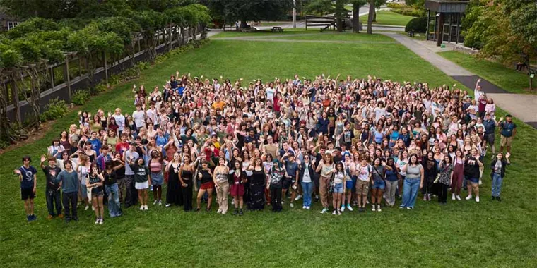 Large group of people gathered on a lawn, some with raised arms, surrounded by trees and pathways.