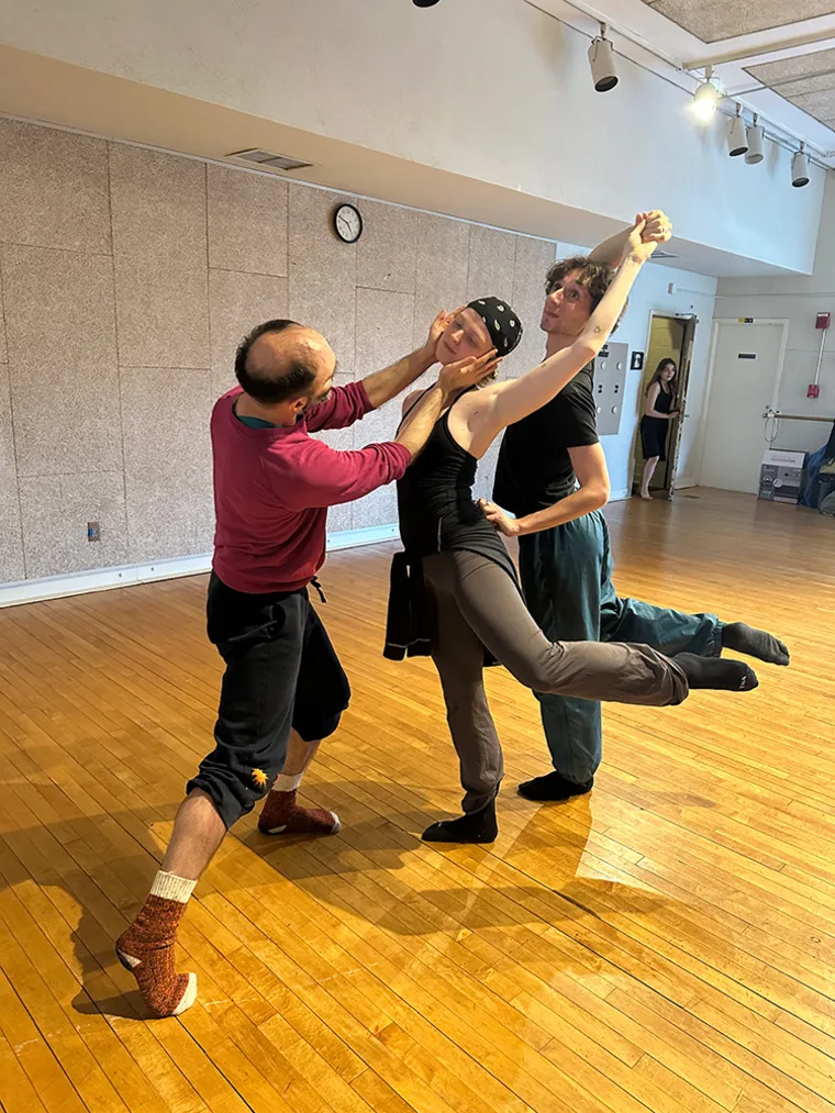 Three people in a dance studio practicing a lift, with wooden floors and lights overhead. A person watches from a doorway.