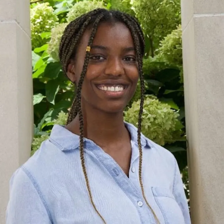Person smiling with braided hair, wearing a light blue shirt. Green leafy background and beige stone pillars.