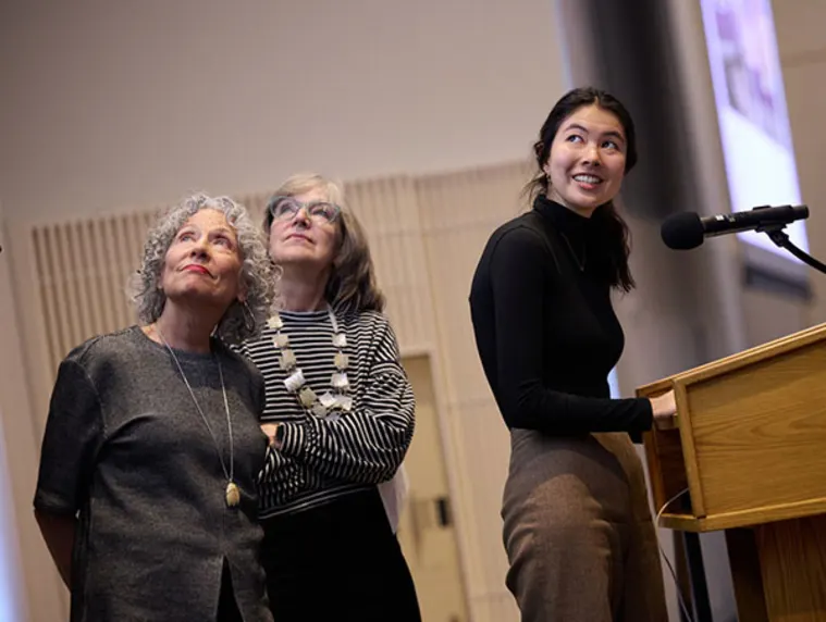 Three people stand near a podium, looking up, with one speaking into a microphone.