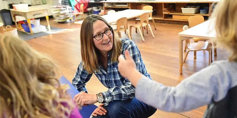 Person kneeling and smiling while interacting with a child in a classroom setting with wooden floors and small tables.