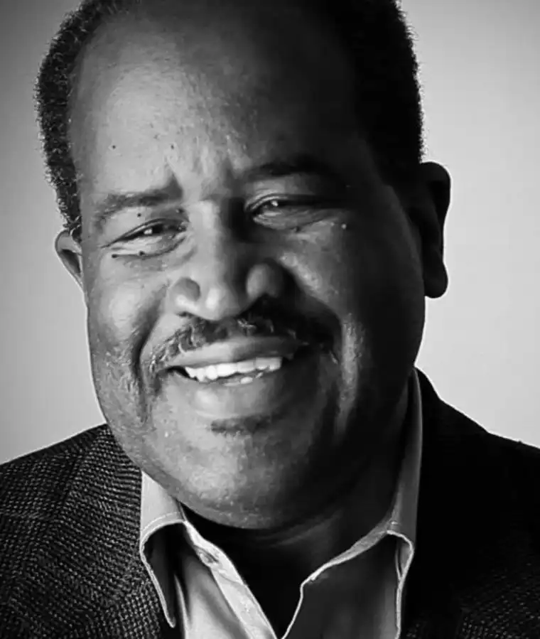Smiling person with short hair and a mustache, wearing a suit jacket and shirt in a black and white portrait.