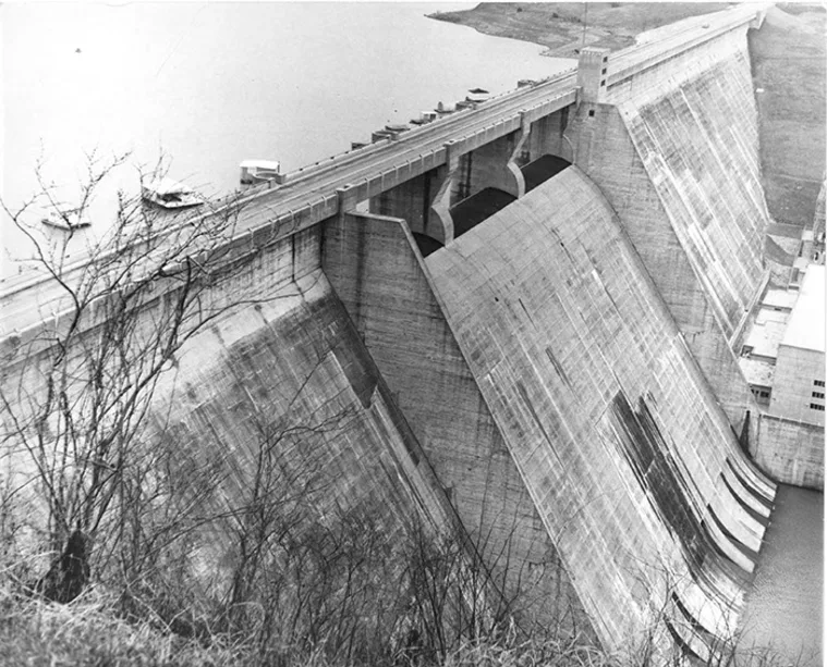 A large, concrete dam with arches is seen from above, holding back a body of water. The surrounding area has sparse foliage.