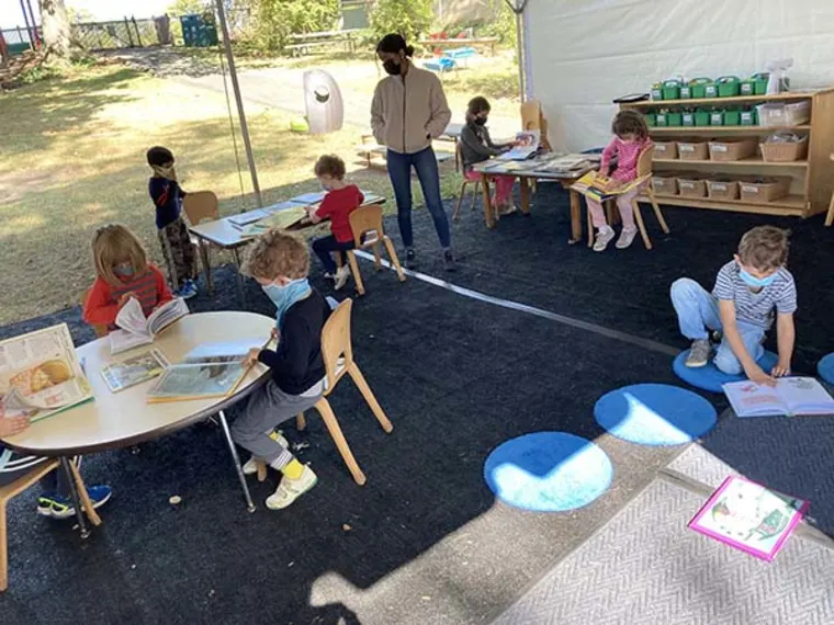 Children sitting and reading books at tables outside; some wearing masks. An adult stands nearby under a canopy.