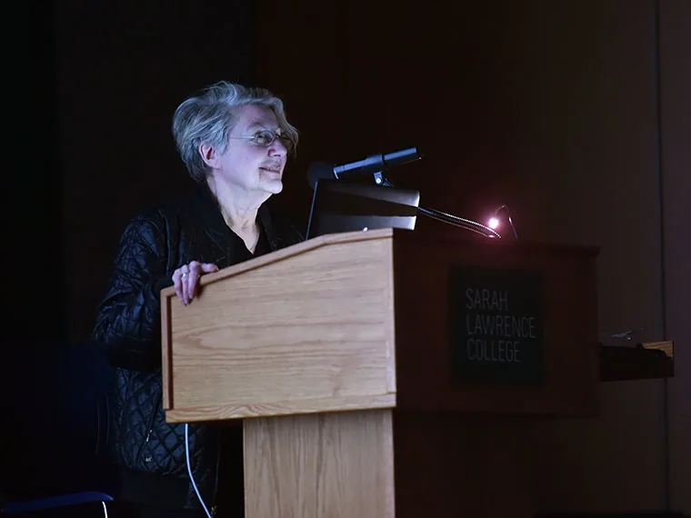 Person speaking at a podium with Sarah Lawrence College logo, illuminated by a laptop and microphone light.