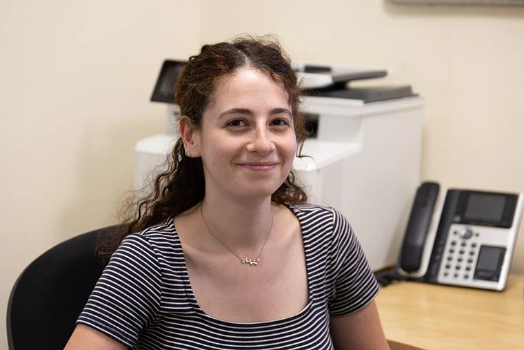 Person sitting at a desk, smiling. A printer and phone are visible in the background.