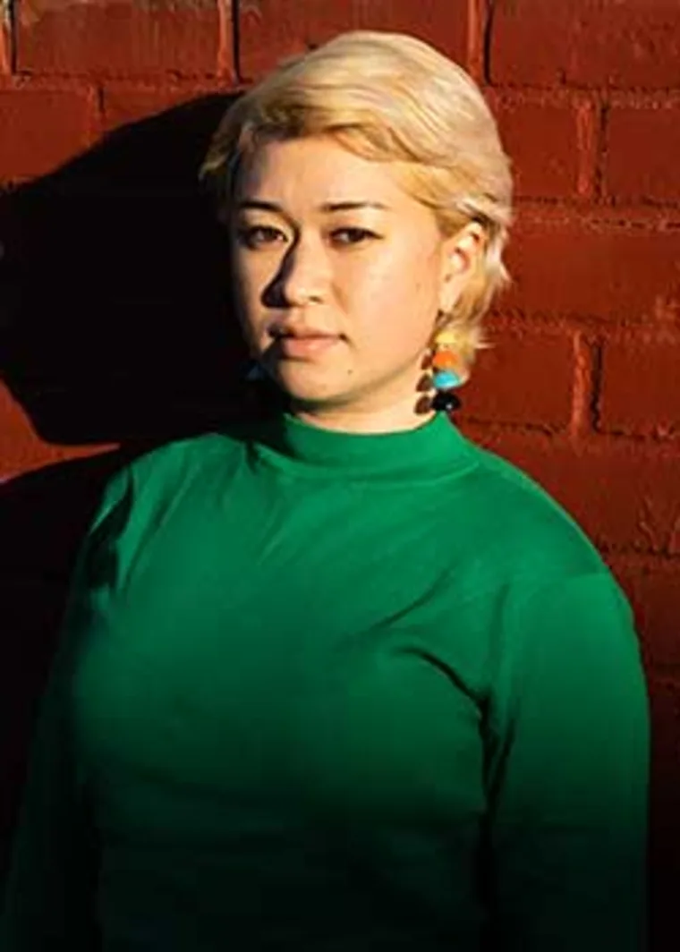Person with short light hair in a green shirt stands against a red brick wall, with strong sunlight creating a shadow.