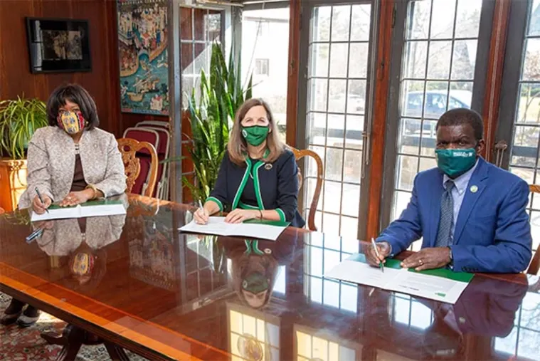 Three people wearing masks sit at a table with papers and pens, inside a room with large windows and plants.