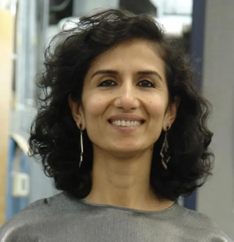 Person with curly hair, smiling, wearing a gray top and dangling earrings, standing indoors.