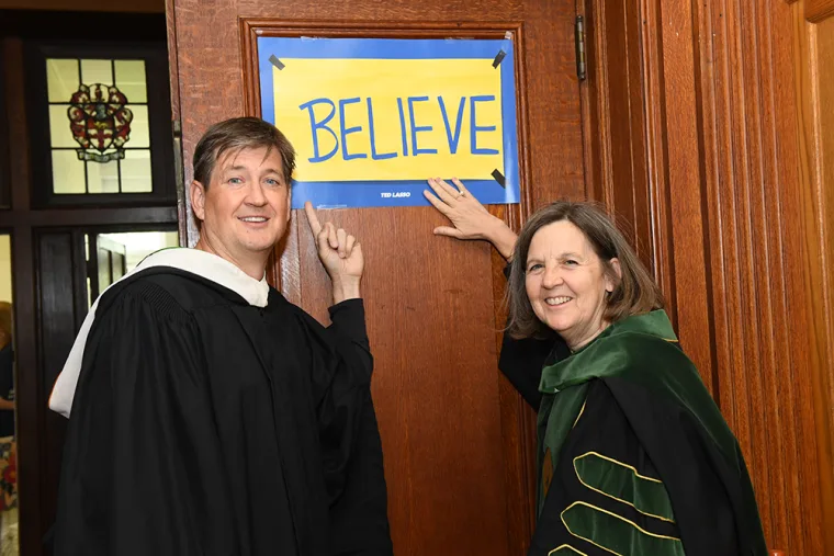 Two people in academic gowns stand by a wooden door with a sign that reads "BELIEVE" in blue letters on a yellow background.