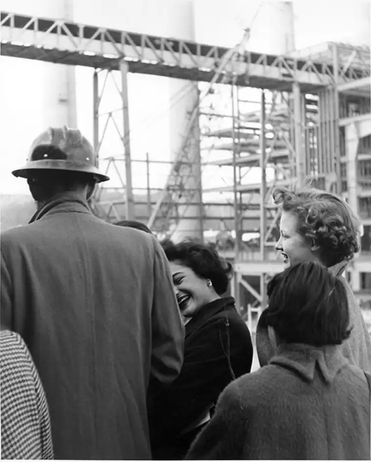 Group of people conversing and smiling at a construction site with cranes and steel beams in the background.
