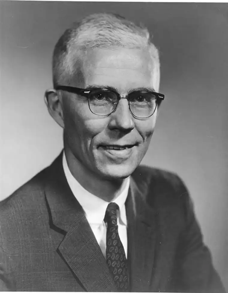 Person with short hair, glasses, and a suit in a black and white portrait, smiling slightly at the camera.