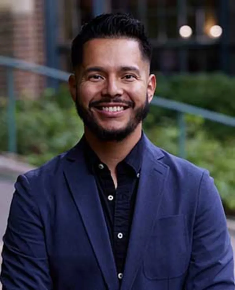 Person with short hair and beard, wearing a dark blazer and smiling, stands outdoors with greenery and a building in the background.